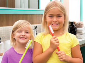 Two young girls in the bathroom holding giant toothbrushes.
